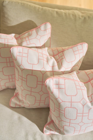 Three Pink Lattice Pillows with a geometric design and pink piping are arranged on a beige couch, illuminated by natural sunlight.