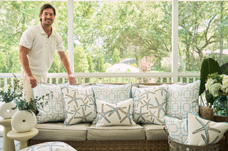 A smiling man stands beside an outdoor wicker sofa adorned with coastal white and blue pillows, including the Starfish Oversized Pillow, on a bright porch surrounded by lush greenery and potted plants.