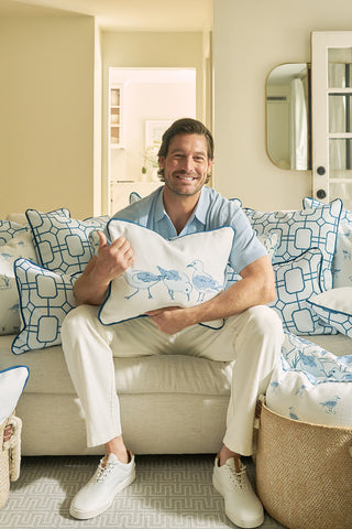 A man sits on a cream sofa, smiling and giving a thumbs up while holding a Sandpiper Trio with Blue Lattice Lumbar Pillow. The sofas blue and white patterned pillows enhance the cozy, beachy vibe of the neutral room.