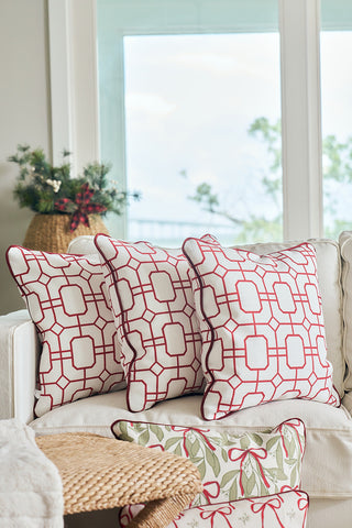 Three white throw pillows, including a Red Holiday Lattice Oversized Pillow with a bold geometric pattern, are arranged on a cream sofa. A large window with a blurred outdoor view and greenery in a woven basket is behind them.