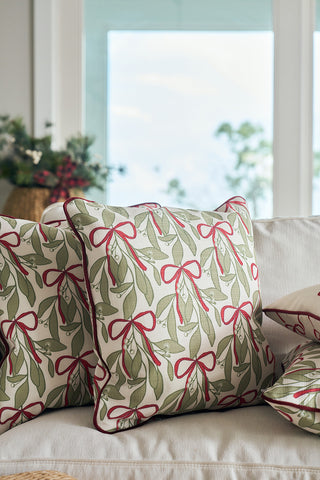 Two decorative holiday pillows, including the Oh Mistletoe Oversized Pillow, sit on a light sofa. The Oh Mistletoe pillow features an olive green leaves and red bow pattern. Blurred windows and festive greenery are in the background.