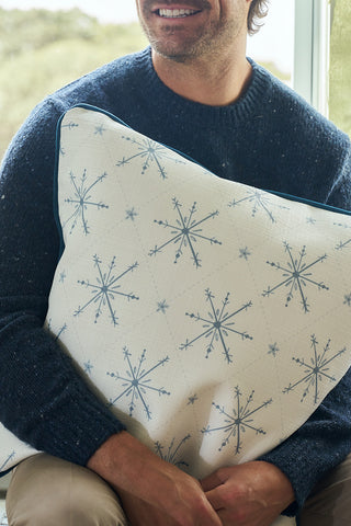 A man in a dark blue sweater holds the Luxe Snowflakes Oversized Pillow, its festive blue design from Sewing Down South brightening his indoor space as he sits smiling by the window.