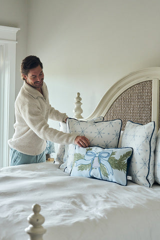 A man in a cream sweater arranges a Holiday Swag Extended Lumbar Pillow and a blue Christmas pillow on a neatly made bed in a bright, cozy bedroom with neutral walls and large windows.