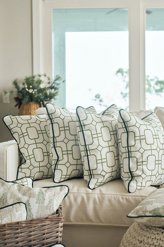 A beige sofa with three white throw pillows, one being the Green Holiday Lattice Oversized Pillow, sits by large windows. A nearby basket of patterned cushions and a potted plant complete the cozy holiday ambiance.