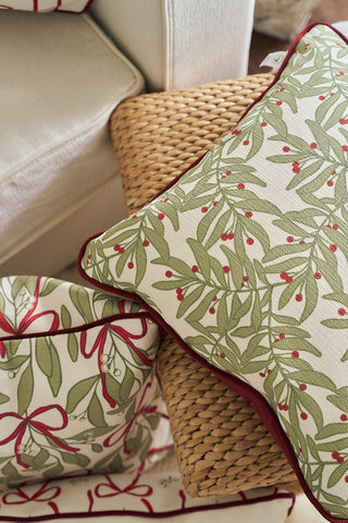 A Jolly Holly Pillow and a fabric basket with green leafy and red berry patterns are arranged on a woven rattan seat next to a white upholstered chair.