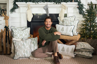 A man in a green sweater sits on the floor by a fireplace, surrounded by holiday pillows including the Green Holiday Lattice Oversized Pillow. The mantle is decorated with stockings and festive garland.