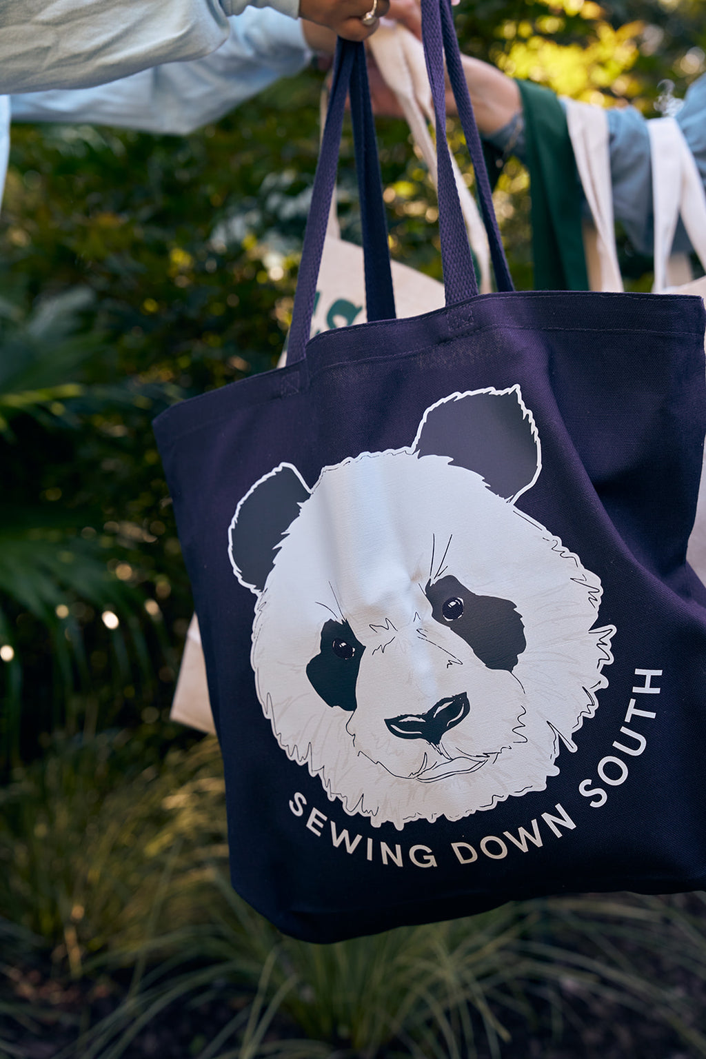 A person holds a navy blue Panda Tote Bag featuring a panda face and Sewing Down South text, with a background resembling a UFO landing site surrounded by lush green foliage, suggesting an outdoor setting.