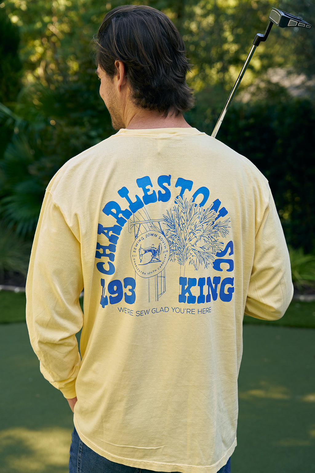 On a sunny day near the Charleston Storefront on King Street, a woman wearing SDS King Street Long Sleeve Tee and ripped jeans putts a golf ball on a small green. A man in the same tee crouches nearby, surrounded by trees and flowers.