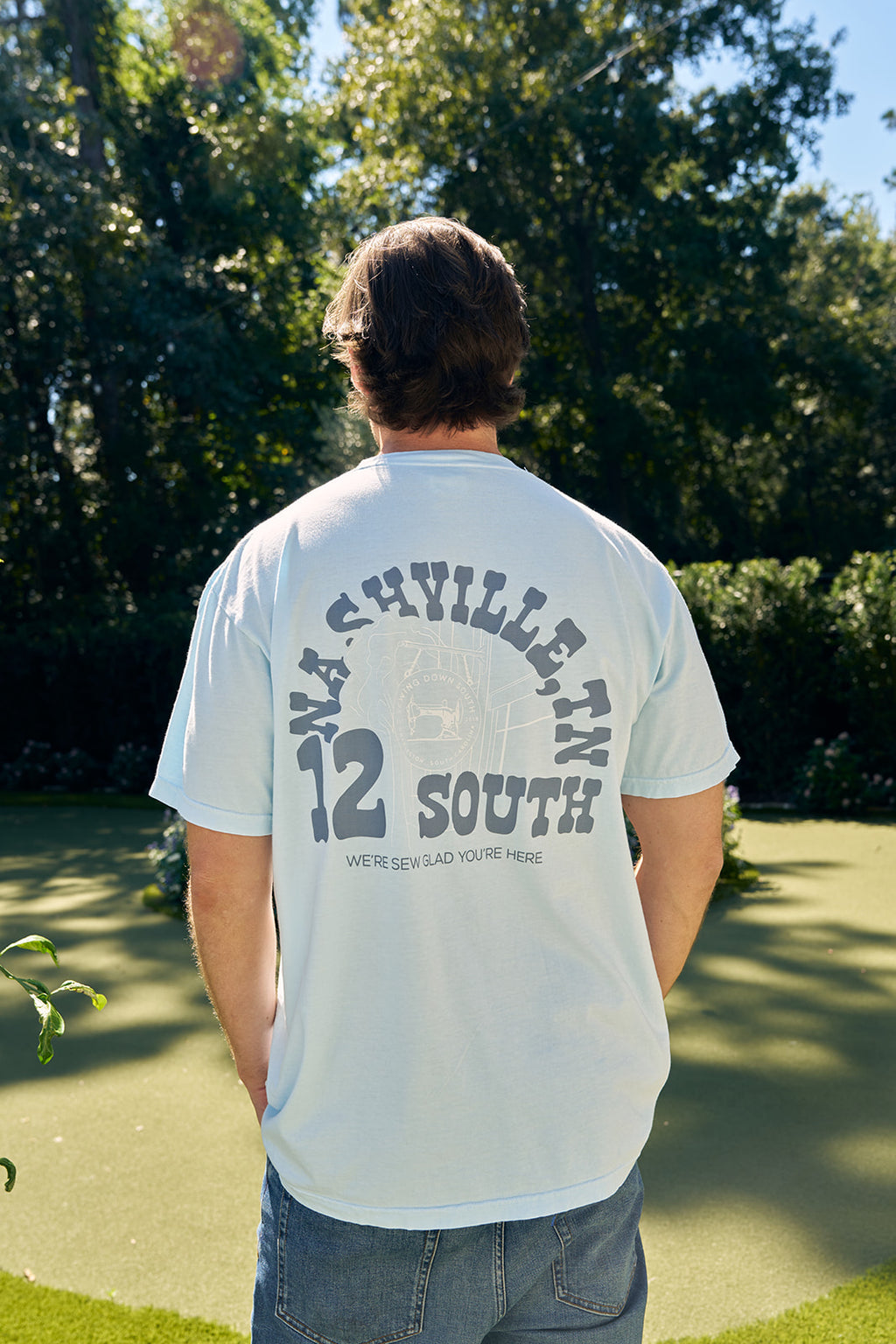 A man strolls a stone path in casual attire, wearing the SDS Nashville 12 South T Shirt with a back text. Hes got green pants, a black cap, and holds a drink container. With his right hand raised showing two fingers, he walks down a street lined with parked cars and buildings.