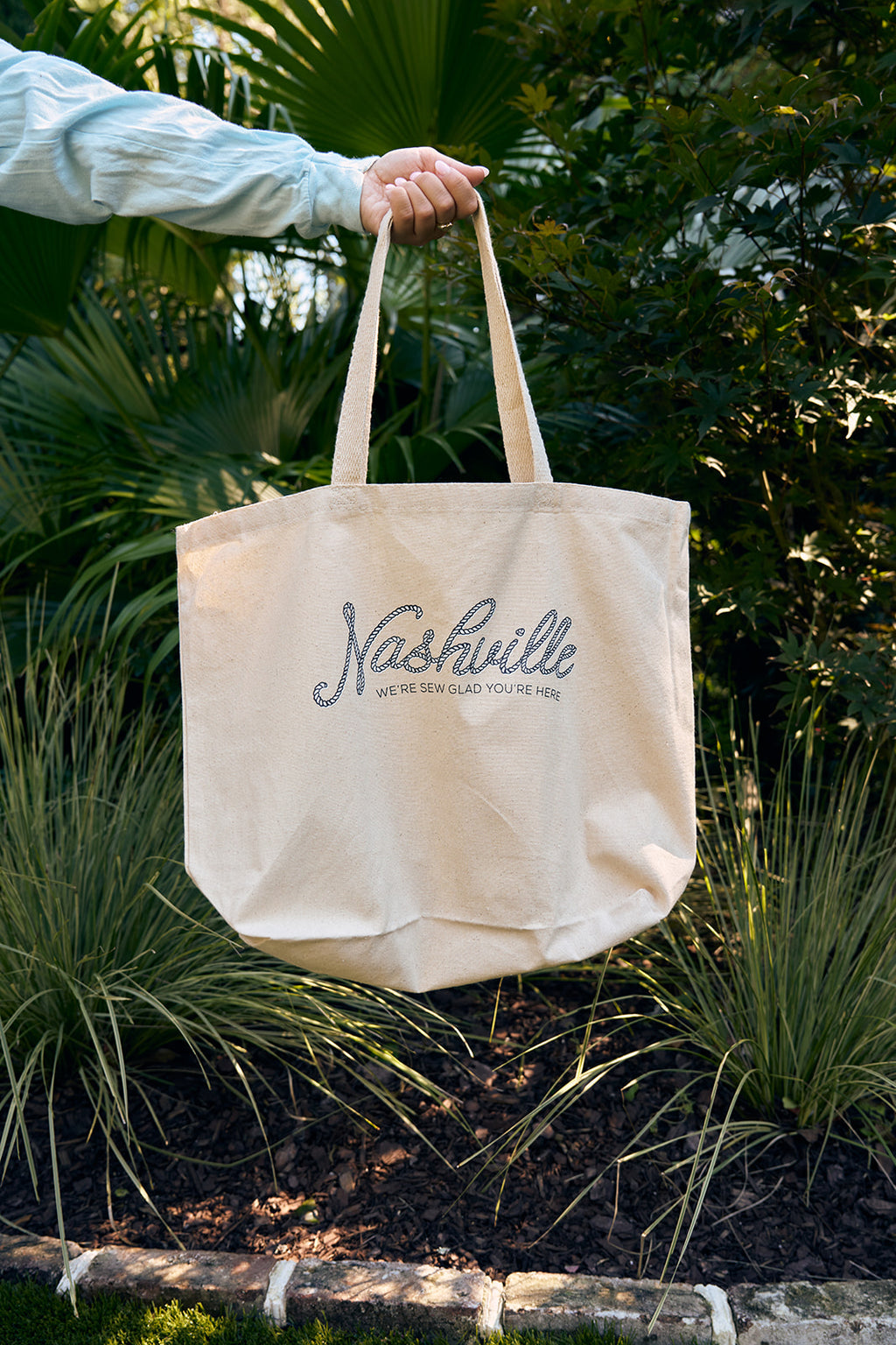 A curly blonde-haired person in a navy sweater holds an SDS Nashville Rope Tote Bag made of heavy cotton canvas while standing in a garden adorned with green plants and pink flowers.