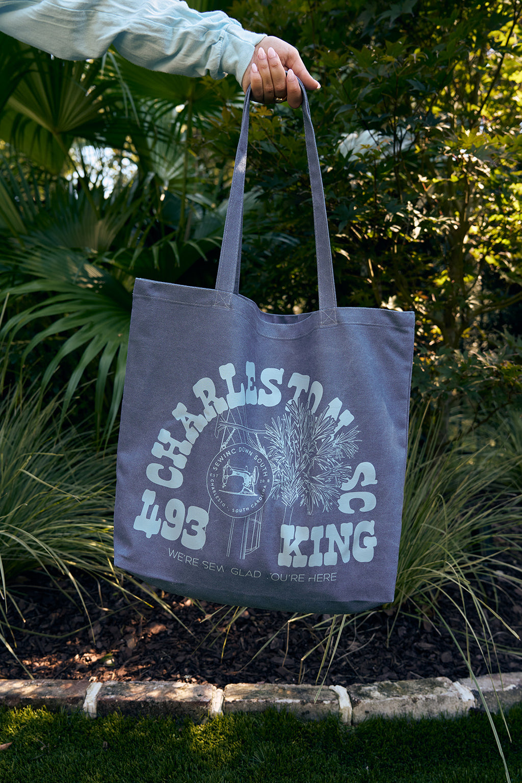 A smiling woman with curly hair carries an SDS King Street Tote Bag—100% cotton canvas, featuring green CHARLESTON SC text—over her shoulder by a pool, next to a friend. Both wear casual green shirts.