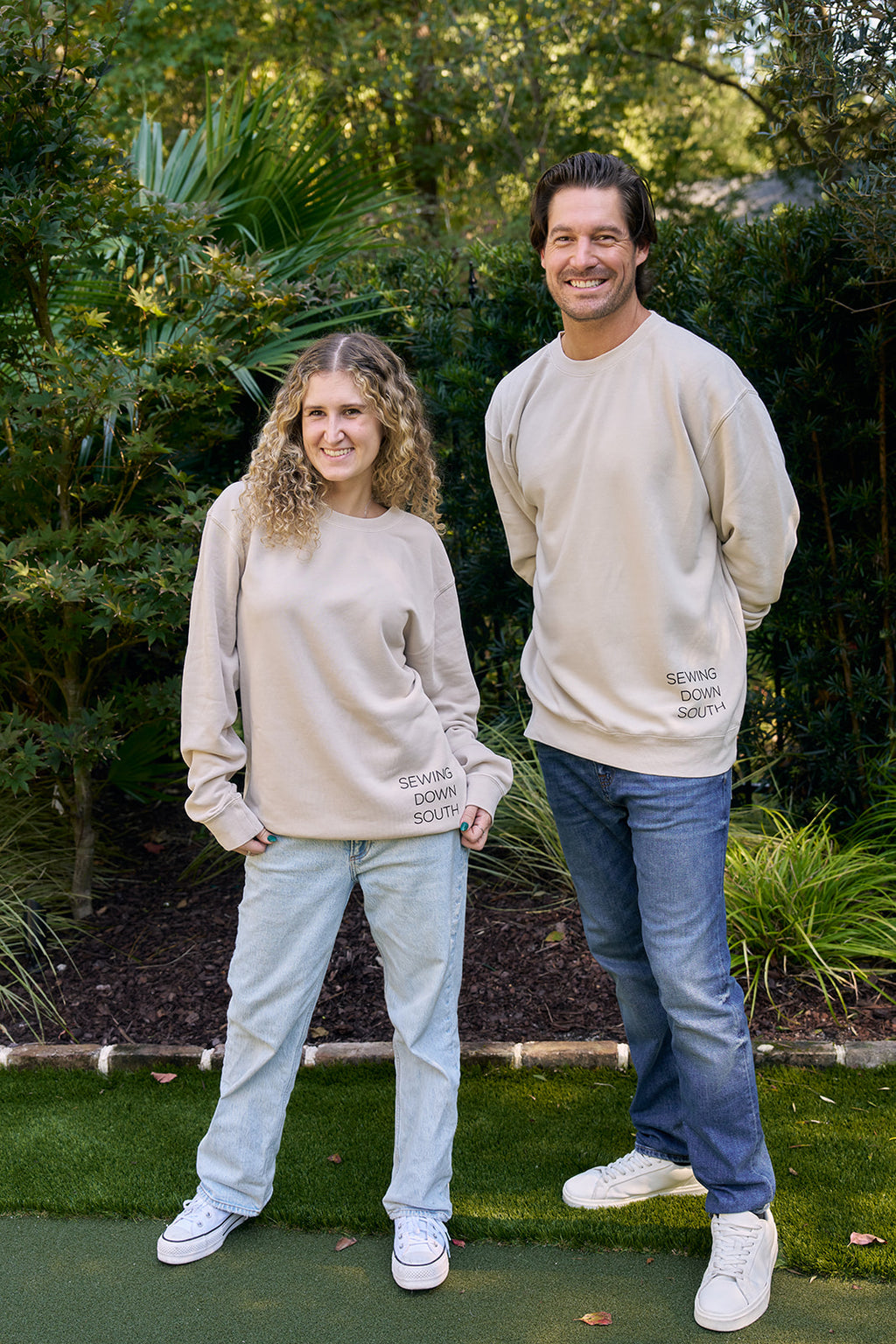 A man stands outdoors facing away, wearing the SDS King Street Crewneck Sweatshirt in Bone. The back features CHARLESTON CO. 193 KIN, plant and sun graphics, and WERE SEW GLAD YOURE HERE beneath the design.