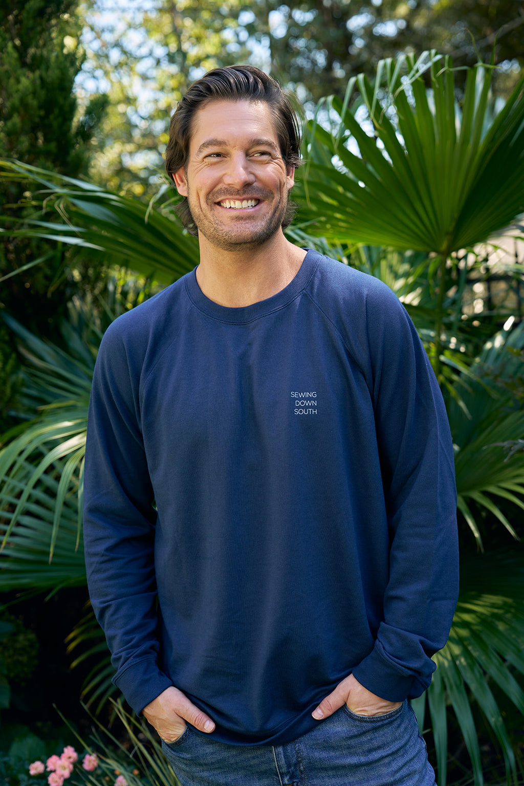 Two casually dressed people outdoors: the left person has curly hair, wearing a navy sweatshirt with Charleston King from the King Street Crew collection. The smiling person on the right wears an Indigo SDS King Street Crewneck Sweatshirt with Goodbye Good Vibes on the front.