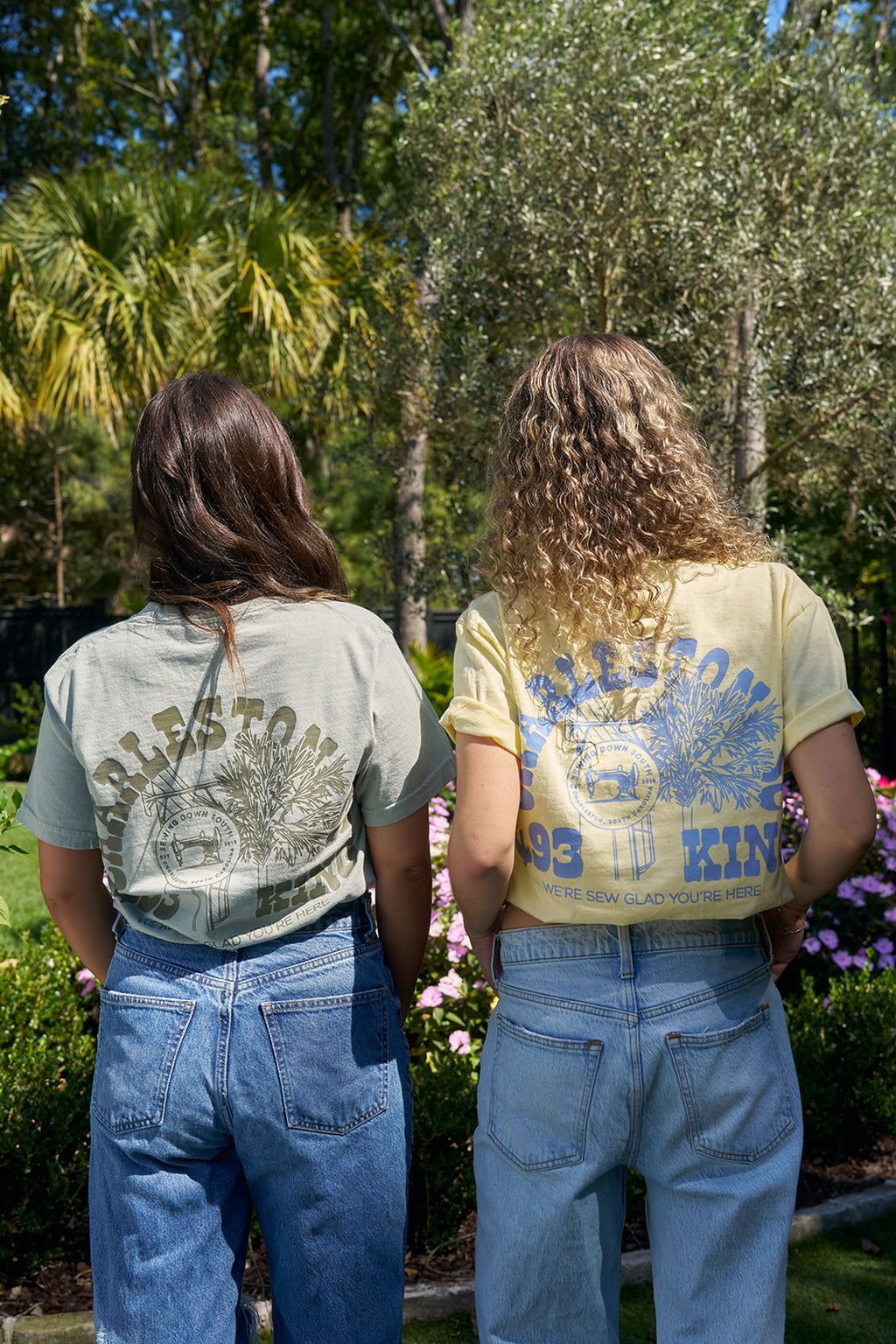 A person with brown hair stands in a garden of pink flowers and palm trees, wearing the vibrant 100% ring spun cotton SDS King Street Flagship T-Shirt. The shirt features a clock tower design, CHARLESTON, KING, and celebrates your visit to this charming Charleston storefront.