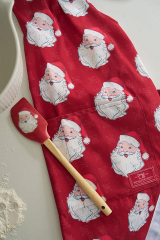 A red apron decorated with Santa faces lies on a white surface beside the Santa Spatula, a BPA-free silicone spatula with a wooden handle. Scattered flour hints at baking fun, making this duo an ideal holiday baking gift.