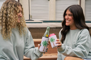 Two women in light green sweatshirts sit outside, smiling and toasting with drinks in white Holiday Palmetto Koozies, available in a 4 PACK (Skinny or Standard) and decorated with palm tree designs.