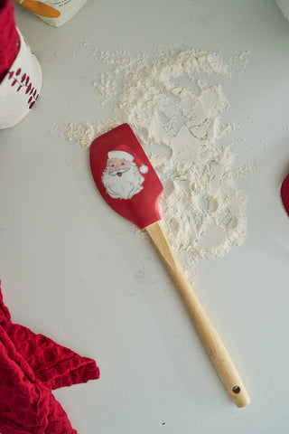 A red BPA-free silicone Santa Spatula with a festive design rests on floured white surface beside a red textured cloth and cookie cutter shapes—perfect as a baking gift.