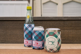 Three beverages—a glass bottle, can, and metal cup—sit on wood, each in a Holiday Plaid Koozie 4 PACK (Skinny or Standard) labeled Bourbon Down South. These neoprene holders keep drinks cool on a wooden surface outdoors.