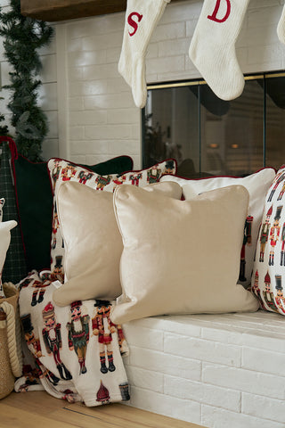 Two beige throw pillows and a velvet pillow rest on a white brick bench next to the Nutcracker Stripe Pillow in Angora; more nutcracker designs sit by the fireplace, which is decorated with white knit stockings and greenery.