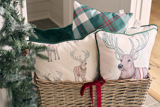 A wicker basket with a red ribbon holds three Christmas pillows—including the Green & Red Plaid Lumbar Pillow—next to a pine garland adorned with pinecones.