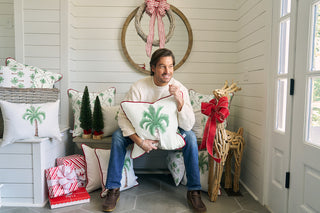 A man in a white sweater and jeans sits on a bench holding an Oversized Holiday Palmetto Pillow, surrounded by festive pillows, small trees, a wicker reindeer with a red bow, and wrapped gifts in a bright, cozy entryway.