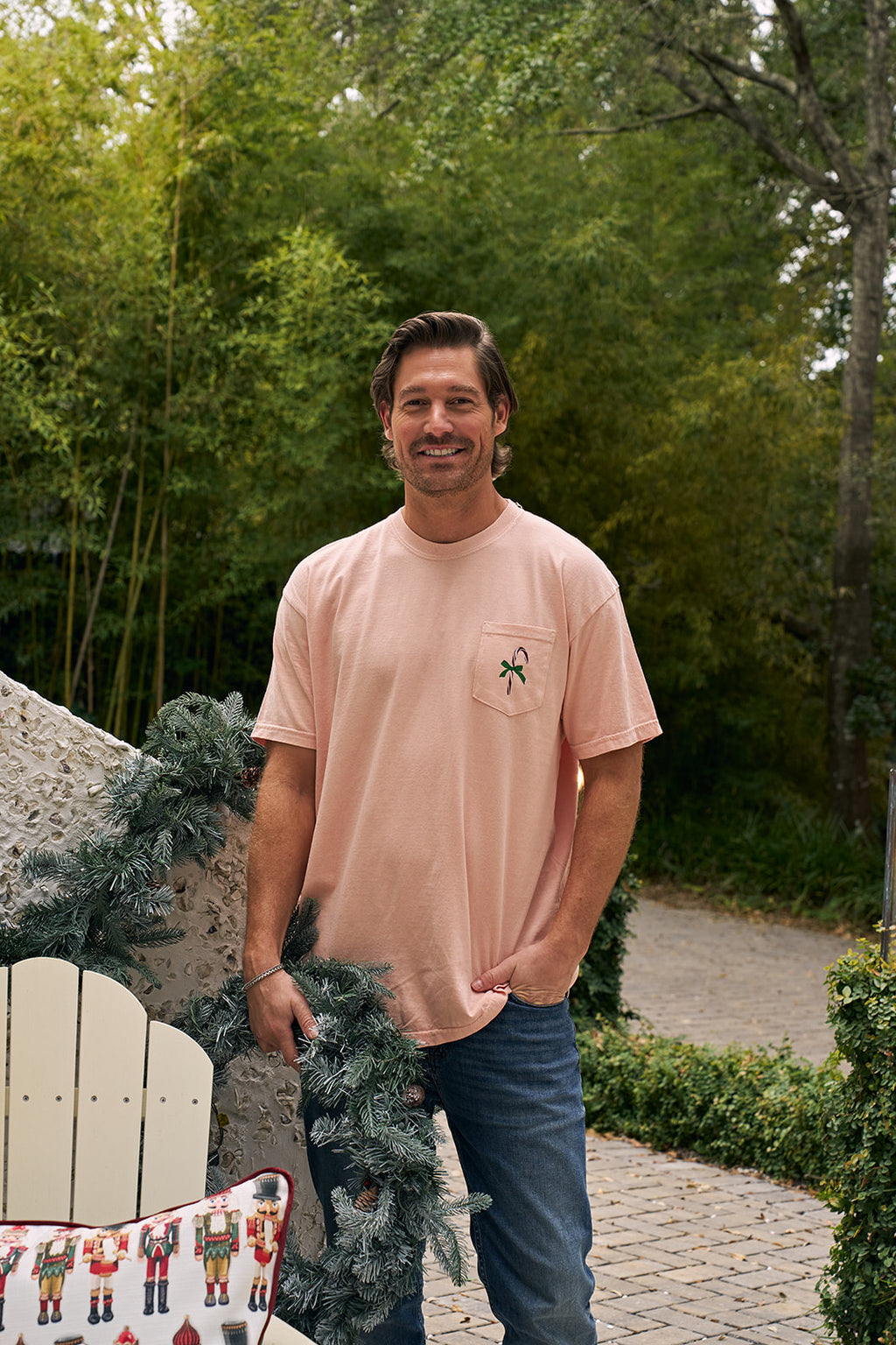 A person wearing the Pastel Nutcracker T-shirt, featuring three colorful nutcracker designs on the back, stands outdoors near a decorated garland with greenery in the background—a festive holiday apparel choice.