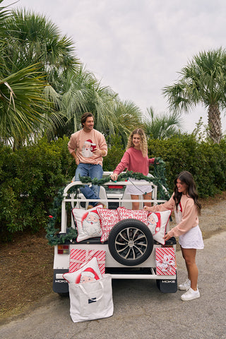 Three people pose by a white vehicle decorated with Christmas pillows and greenery. Two, wearing Santa Crewneck sweaters, sit on the back while one stands nearby. Palm trees and a cloudy sky complete this festive scene.