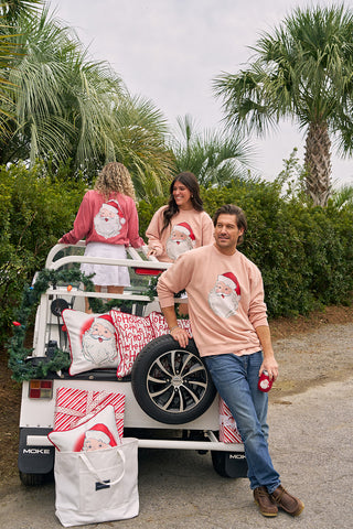 Three adults wearing Santa Crewneck sweatshirts pose by a white golf cart decorated with Christmas pillows and wrapped gifts, set against a backdrop of palm trees and greenery.
