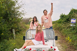 Three people wearing Santa Crewnecks sit and stand on a white open-top vehicle on a dirt path surrounded by greenery. One person raises an arm cheerfully while the others smile at the camera.