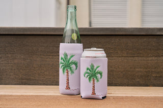 A clear glass bottle and a can, each in a light purple Holiday Palmetto Koozie 4 PACK (Skinny or Standard) featuring a palm tree design, sit on a wooden surface in front of a brown wooden fence.