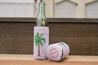 Two Holiday Palmetto Koozies (4 PACK, Skinny or Standard) are shown on a wooden surface—one upright with a glass bottle and the other lying sideways—both featuring palm tree prints against a blurred background.