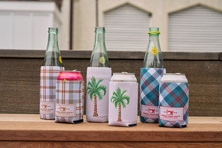 Three glass bottles and three cans with colorful neoprene drink holders, including the Holiday Harvest Plaid Koozie 4 PACK (Skinny or Standard), are arranged outdoors on a wooden surface in front of a brown fence.