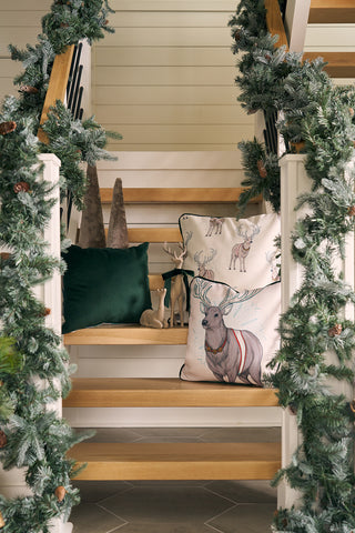 Wooden stairs adorned with pine garlands, the Holiday Reindeer Pillow in a velvet cover, two small deer figurines, and cone-shaped ornaments set a festive holiday scene.