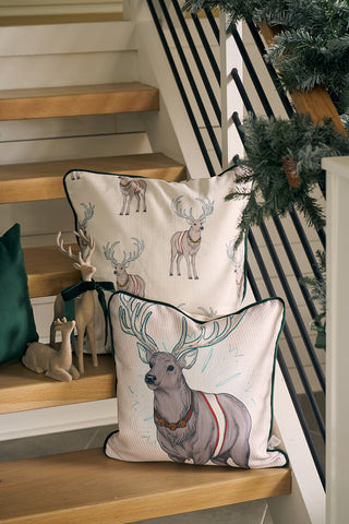 Two Holiday Reindeer Pillows with velvet covers are displayed on wooden stairs beside small reindeer figurines, while a green garland decorates the black stair railing.