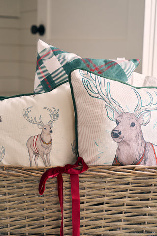A wicker basket holds decorative pillows, including the Holiday Reindeer Pillow, set against a softly lit neutral background with a red ribbon tied to the baskets handle.