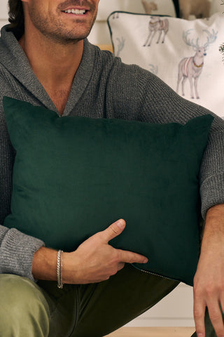 A person in a gray sweater holds a dark green pillow, while in the background, a white pillow with reindeer illustrations and a Green & Red Plaid Lumbar Pillow add to the cozy, festive scene.