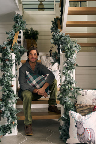 A man sits on a staircase decorated with pine garland, holding a Green & Red Plaid Lumbar Pillow and smiling. Festive reindeer pillows nearby create a cozy holiday atmosphere inspired by Sewing Down South.