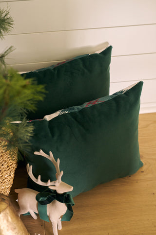 Two dark green throw pillows rest on a wooden floor near a woven basket, pine branches, and a decorative reindeer with a green bow. A Green & Red Plaid Lumbar Pillow completes the cozy, festive look.