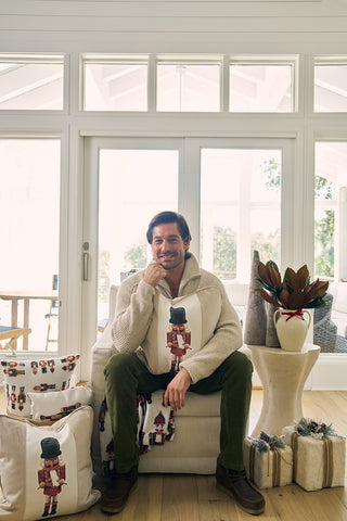 A man sits in a bright, cozy living room holding the Nutcracker Stripe Pillow in Angora. He is surrounded by festive pillows and blankets, with decorative plants and wrapped gifts arranged on a nearby table.