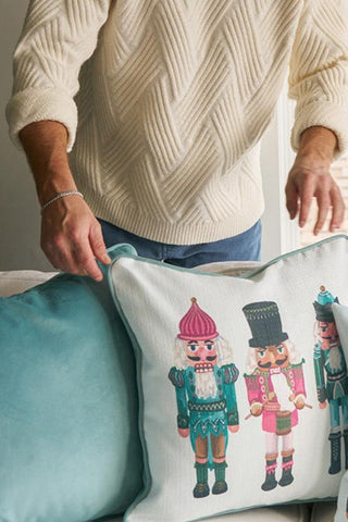 Wearing a cream textured sweater, someone arranges a Pastel Nutcracker Pillow—featuring colorful nutcracker designs—on a light blue couch.