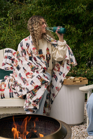 A woman with curly hair relaxes by the fire pit on a white bench, sipping from a bottle wrapped in the Nutcracker Koozie, 4 PACK (Skinny or Standard), and wrapped in a festive blanket, surrounded by greenery.