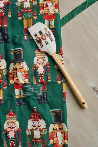 A Nutcracker Apron featuring colorful nutcracker figures and an adjustable neck strap is displayed on a light surface, with a spatula decorated with nutcracker images and a wooden handle resting on top.