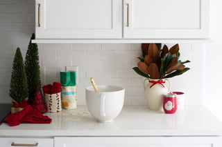 A white kitchen counter features a mixing bowl, spoon, baking ingredients, red towels, Christmas mugs, two small decorative trees, magnolia leaves in a vase, and the BPA-free silicone Santa Spatula against a white tiled backsplash.