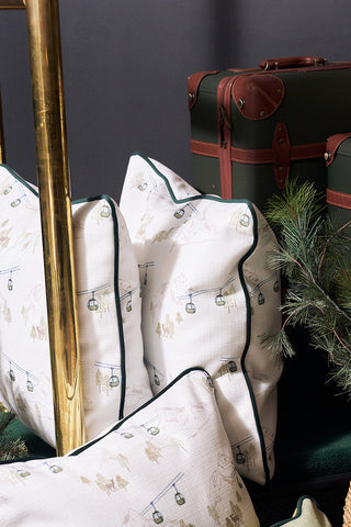 A cozy display featuring the Ski Resort Pillow with après-ski patterns, vintage-style brown suitcases, green pine branches, and a woven basket, all set against a dark backdrop and gold metal frame.