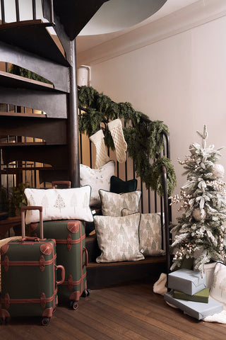 A cozy holiday scene features green and white pillows, including the Row of Trees Extended Lumbar Pillow, on a bench by a spiral staircase decorated with garland and stockings. Nearby are stacked gifts, green vintage suitcases, and a decorated Christmas tree.