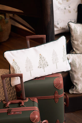 The Row of Trees Extended Lumbar Pillow, a rectangular handmade pillow with embroidered Christmas trees, is displayed on green suitcases with brown leather accents beside a dark wooden staircase in a cozy space.
