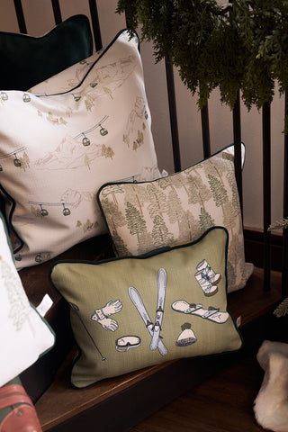 The Winter Sports Gear Lumbar Pillow, ideal for Southern decor, features ski lifts, trees, and ski equipment illustrations on light and green backgrounds—adding a wintry touch to wooden stairs beside a railing and greenery.