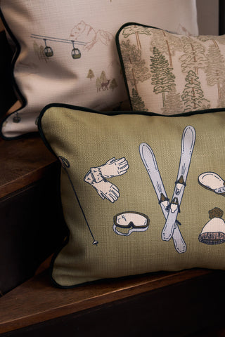 The Winter Sports Gear Lumbar Pillow in olive green with ski equipment illustrations sits on wooden stairs, complemented by a second pillow featuring a modern forest and ski lift design—ideal for adding stylish charm to any room.