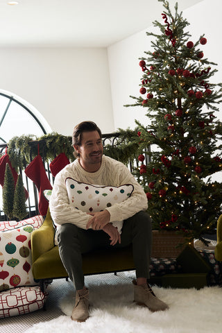 A man in a cream sweater sits on a green chair, holding a Hanging Ornaments Lumbar Pillow. Behind him is a decorated Christmas tree with stockings on a black railing and wrapped gifts on the floor in a bright, cozy room.