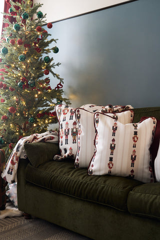 A decorated Christmas tree with lights stands next to a green velvet couch adorned with a cozy Nutcracker Line Up Pillow from Sewing Down South and a matching holiday velvet pillow, all set against a rich blue wall.